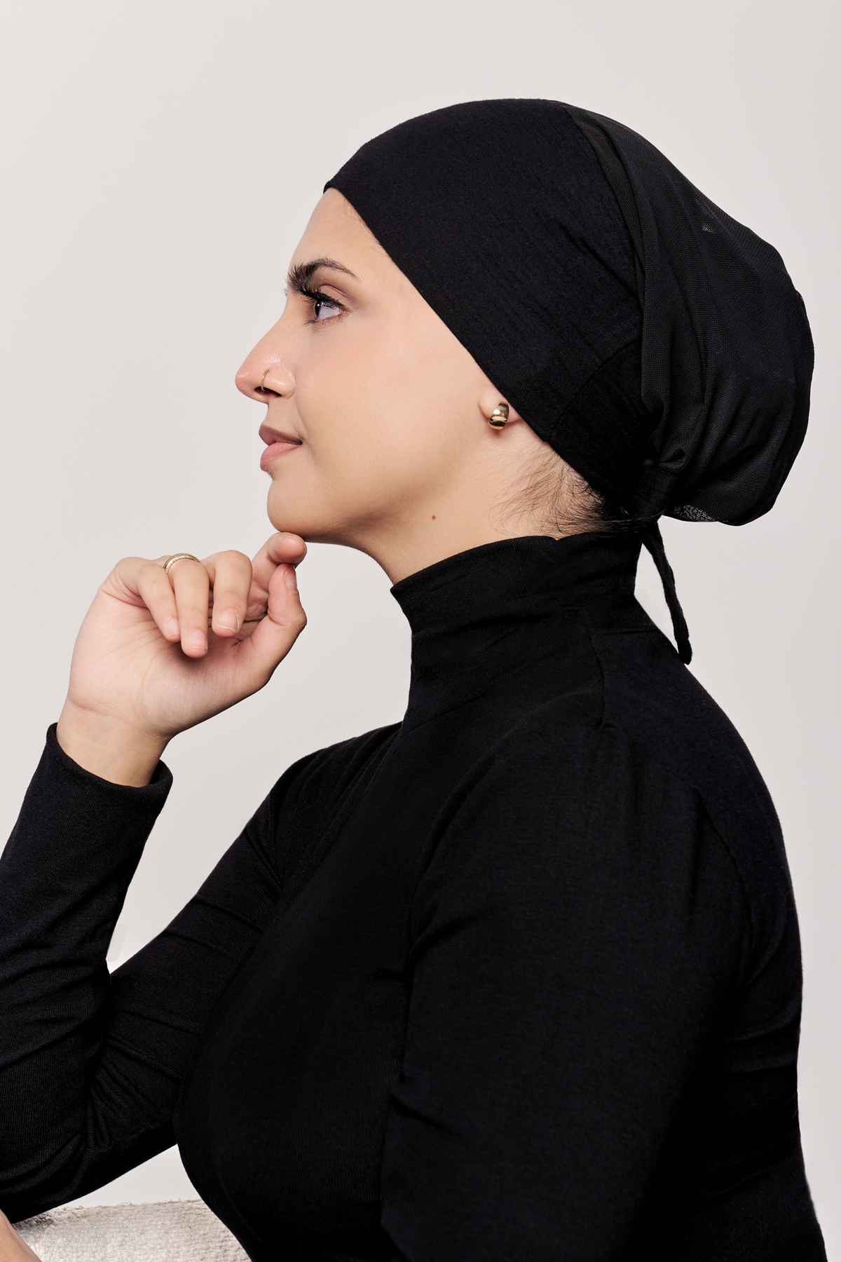 A woman in profile wearing a black turtleneck top and a black underscarf, posed against a light gray studio background.