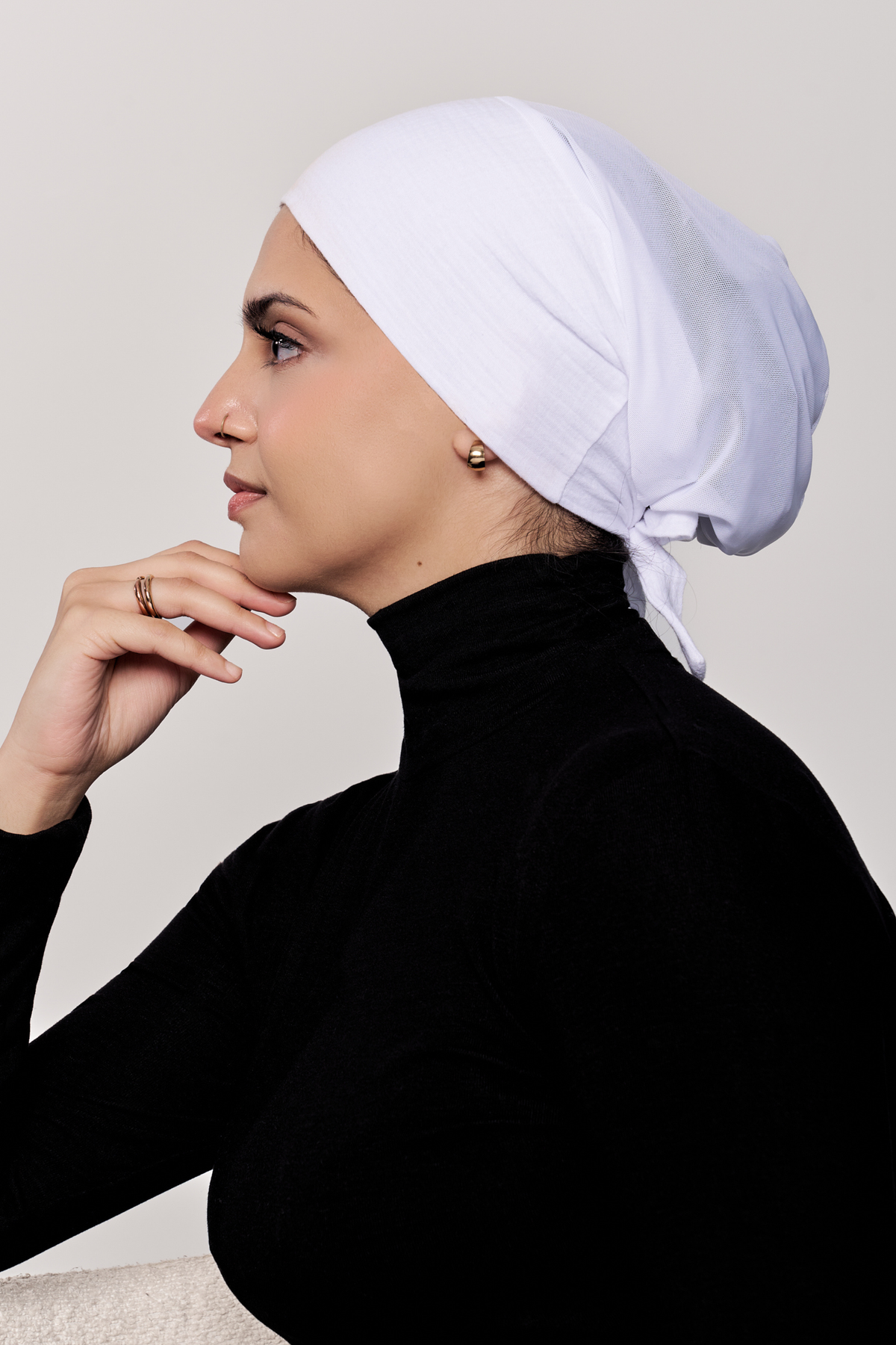 A woman in profile wearing a white underscarf, paired with a black turtleneck top, against a plain light gray background.