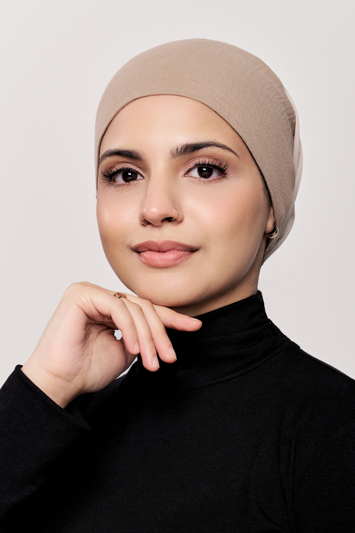 A woman wearing a dark beige underscarf and a black turtleneck top, posing against a light gray studio background.