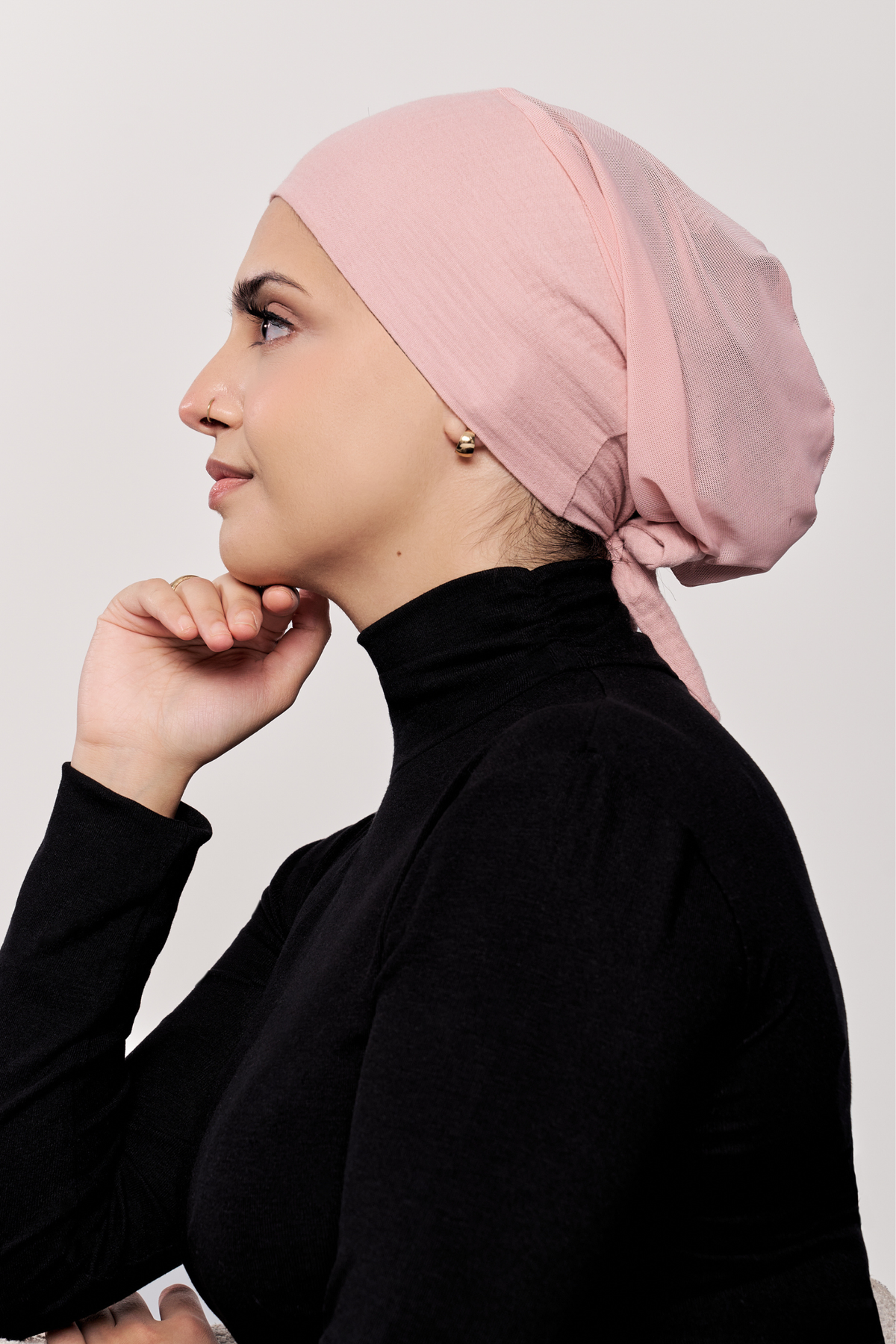A woman in profile wearing a light pink underscarf and a black turtleneck top, photographed against a plain light gray studio background.