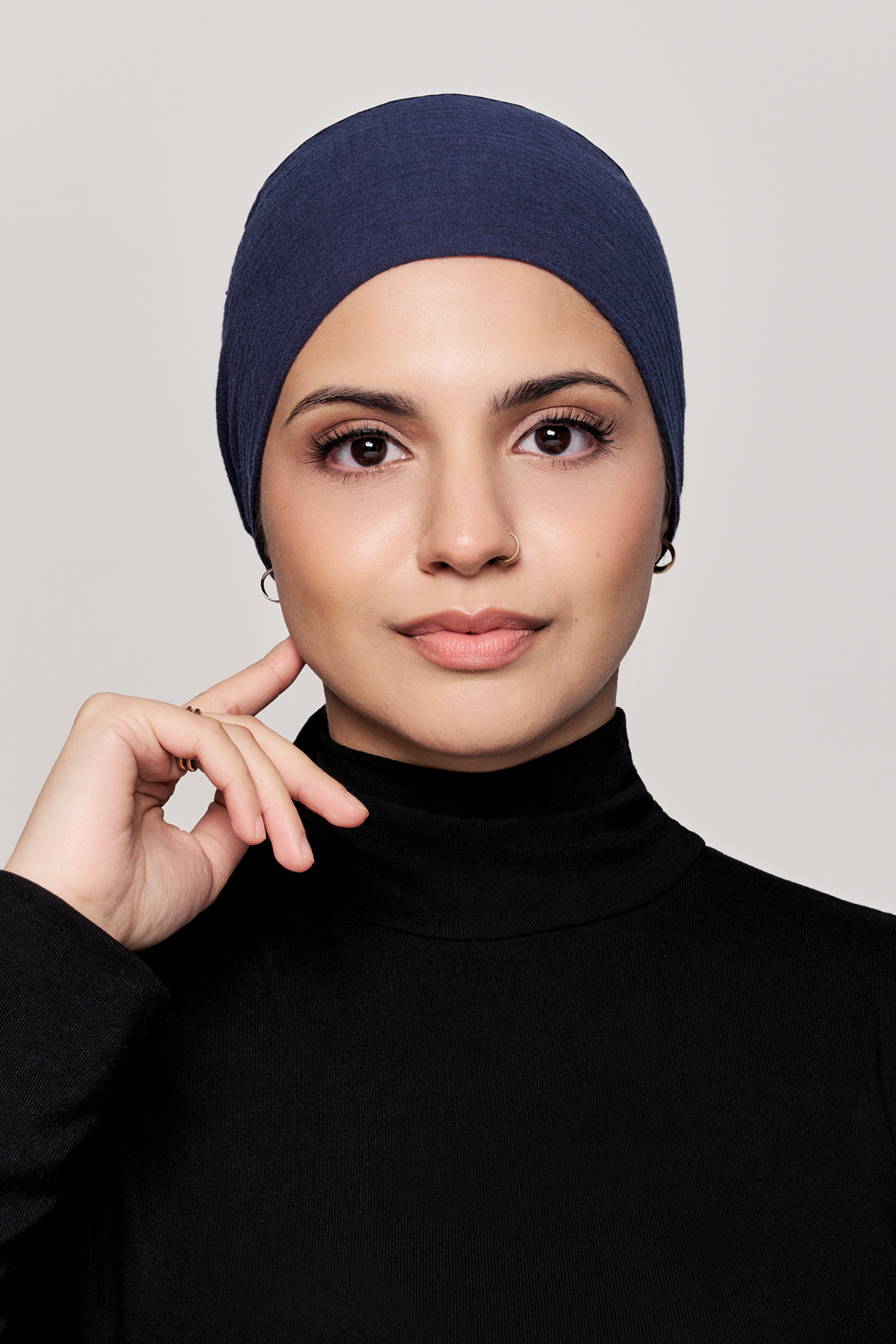 A woman in profile wearing a black turtleneck top and a black underscarf, posed against a light gray studio background.