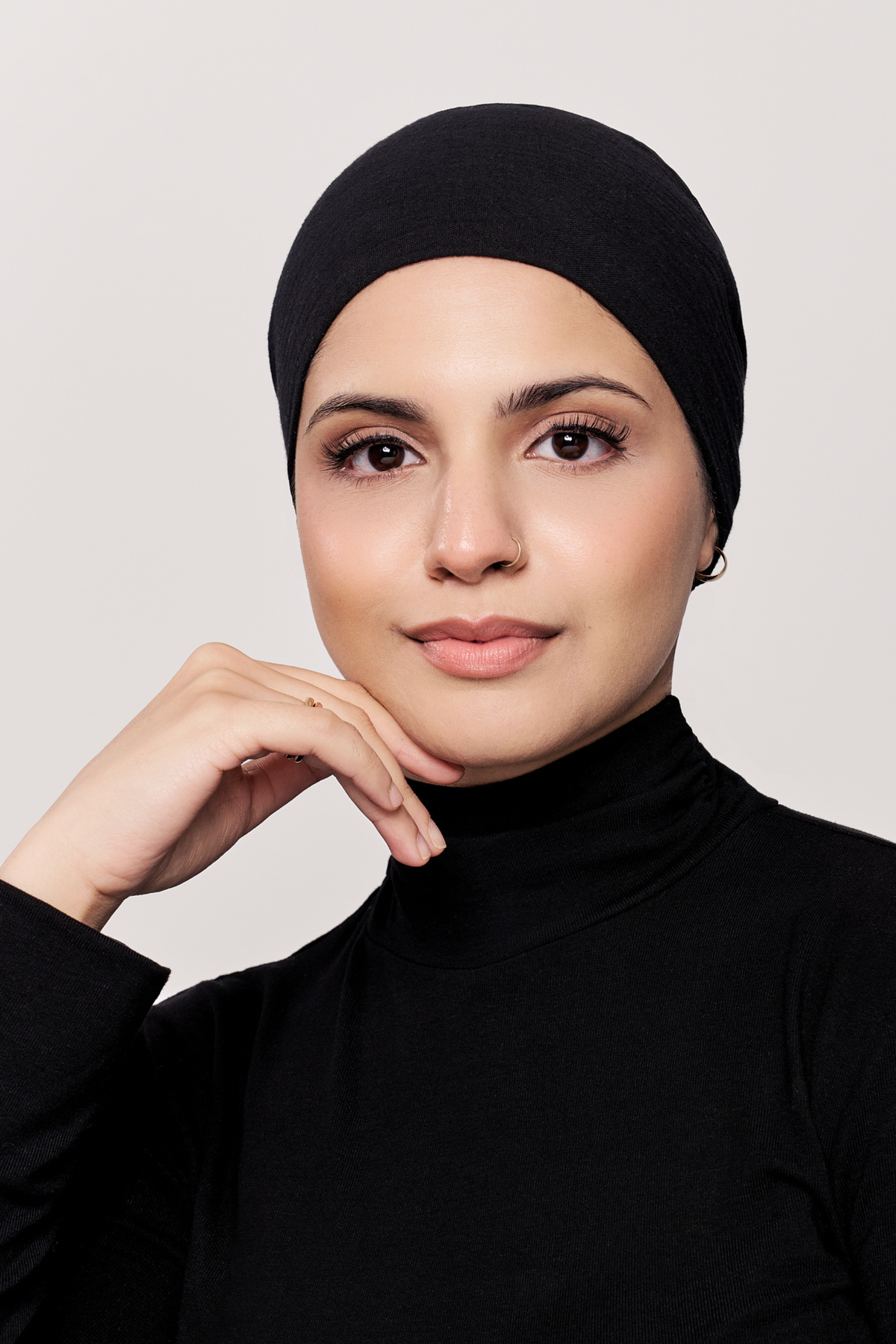 A woman wearing a black underscarf and a black long-sleeve mock-neck top, posing with her hand resting on her chin against a light gray background.