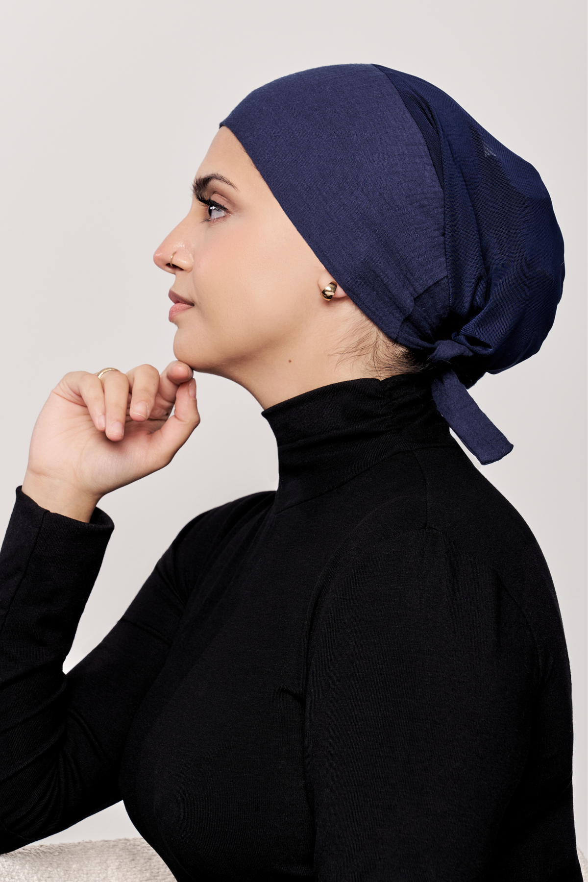 A woman in profile wearing a dark navy underscarf and a black long-sleeve mock-neck top, posed against a light gray studio background.