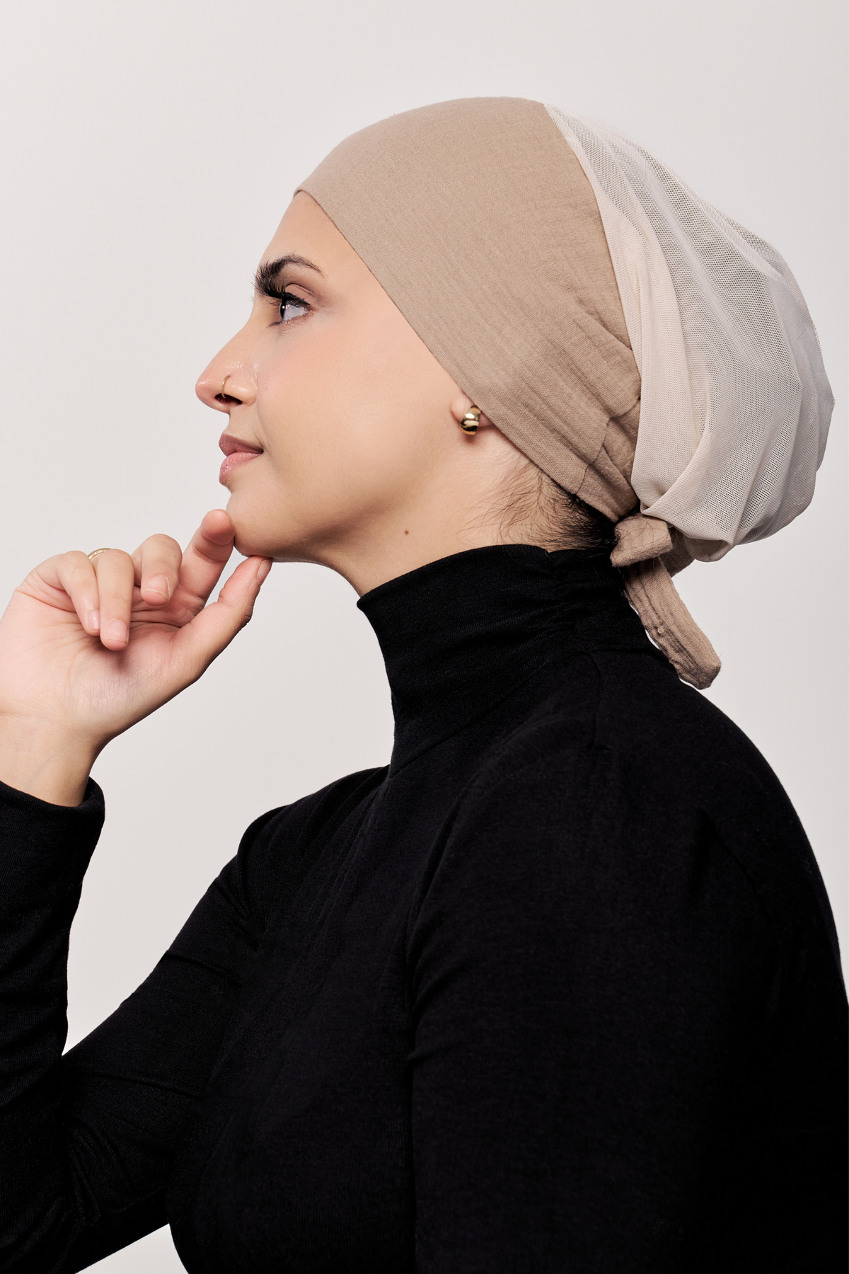 A woman in profile wearing a dark beige underscarf  and a black turtleneck top, posing with her hand near her chin against a plain light gray background.