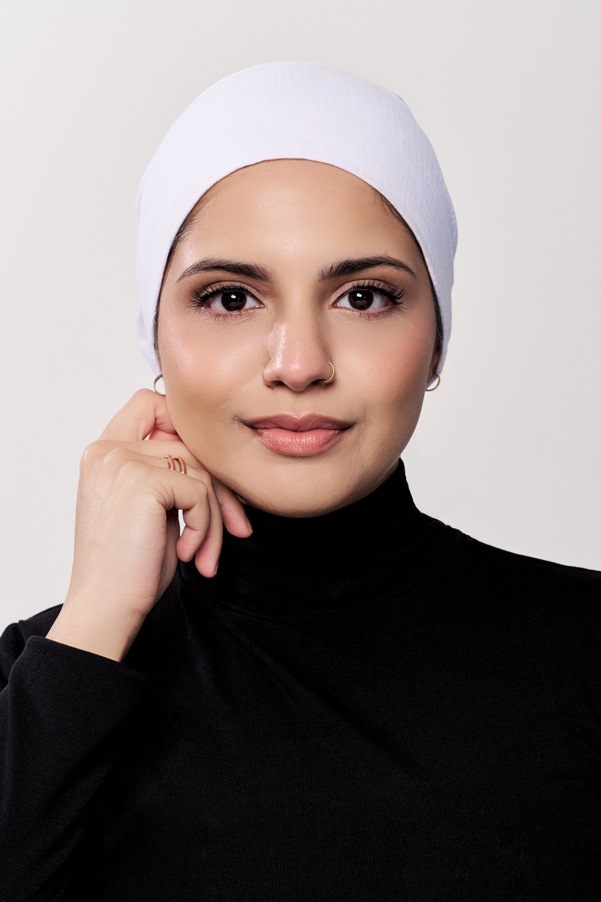 A woman wearing a white underscarf and a black turtleneck sweater, posing against a light gray background.