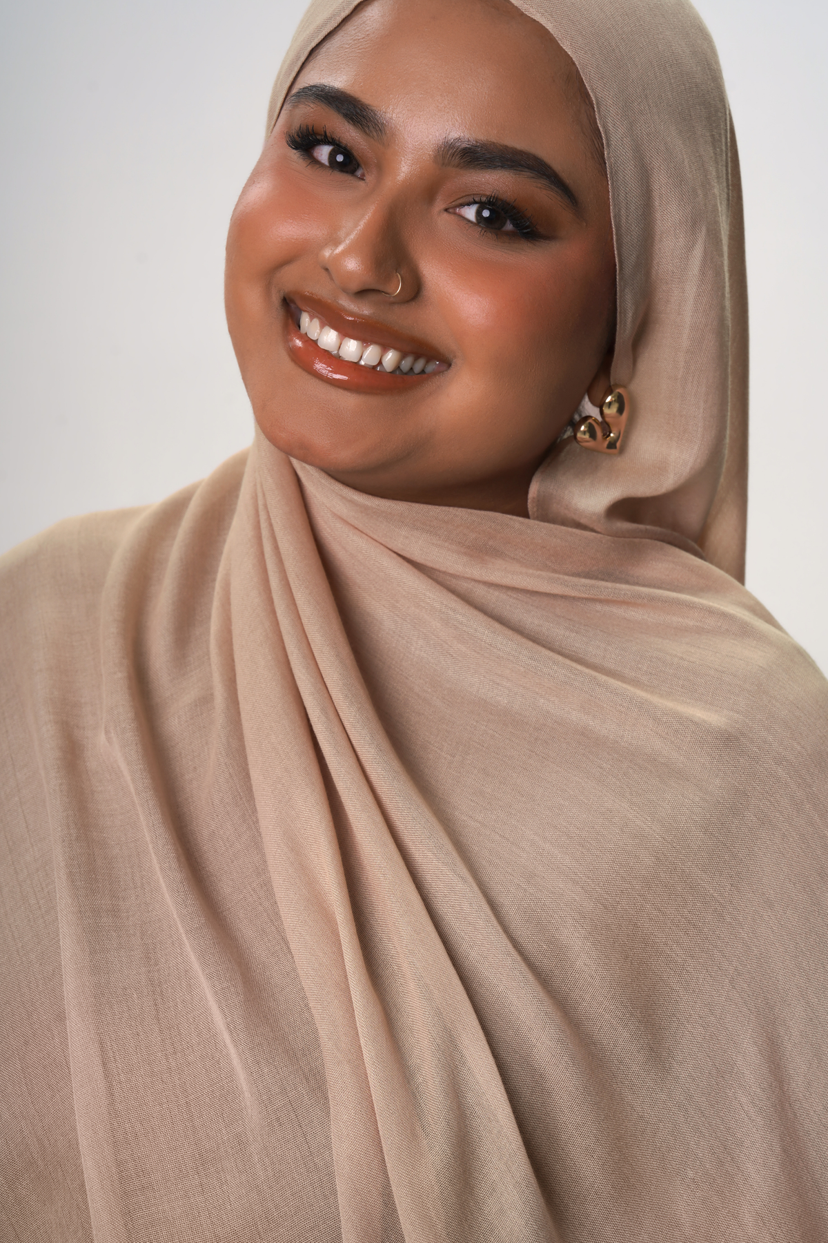 A smiling woman wearing a soft draped hijab in a light taupe-beige color, photographed against a clean light gray studio background.