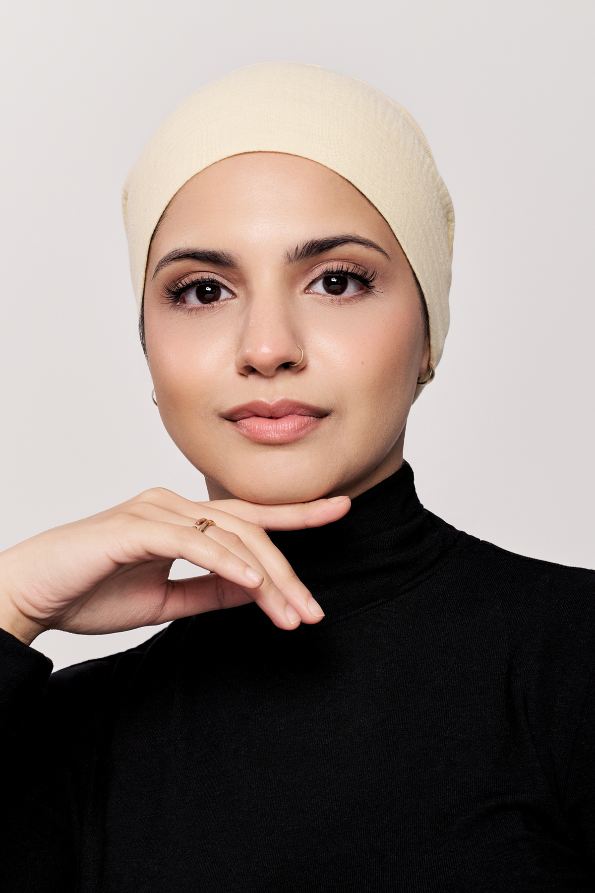 A woman wearing a buttercream underscarf and a black turtleneck sweater, posing against a light gray background.