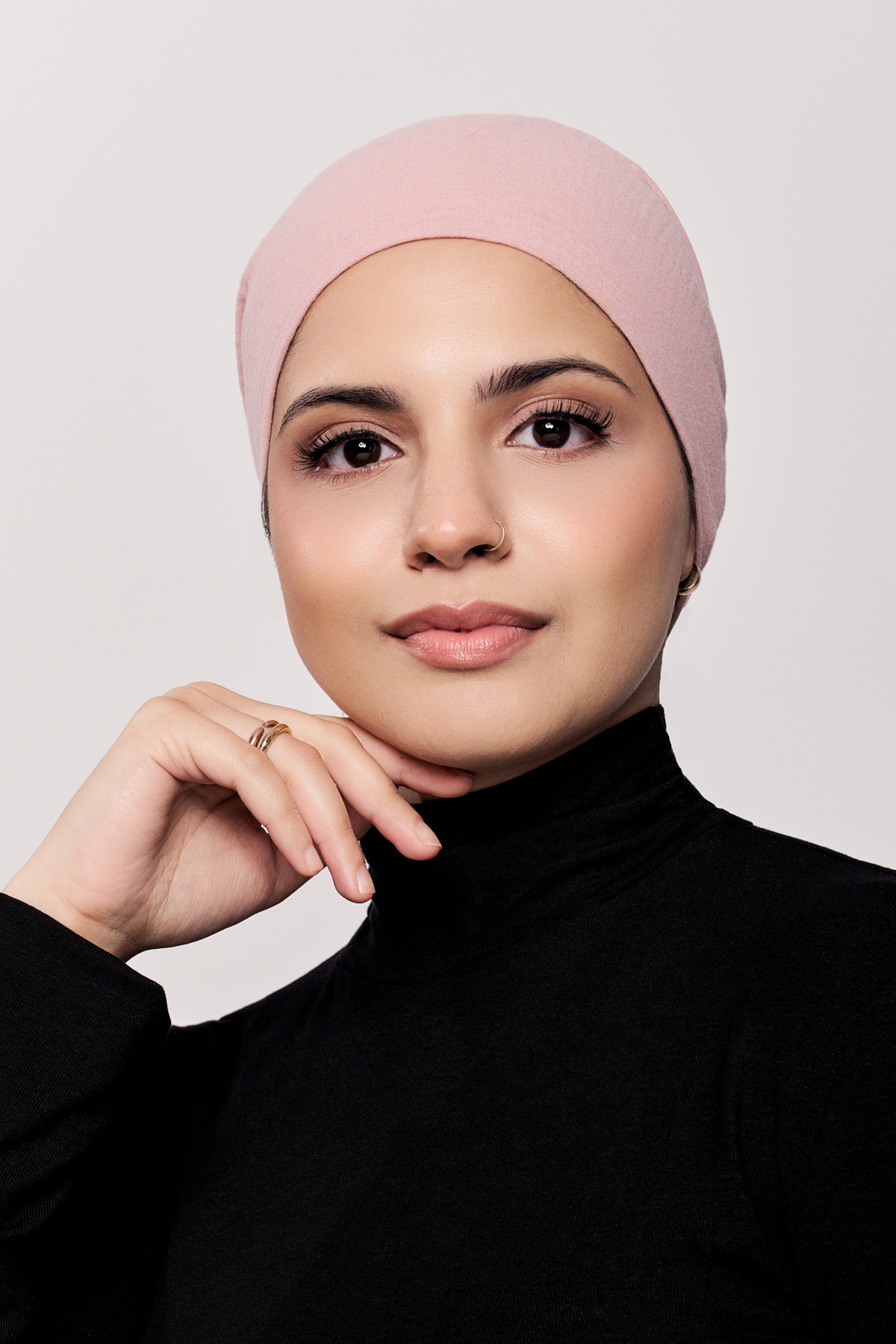 A woman wearing a light pink underscarf and a black turtleneck sweater, posing against a neutral light gray background.