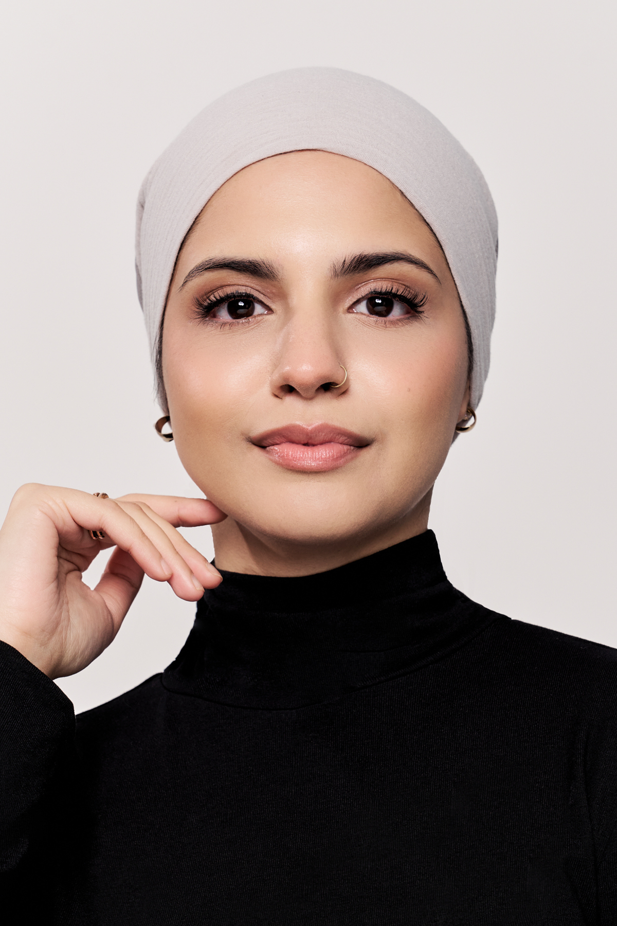 A woman wearing a light gray underscarf and a black turtleneck top, posing against a solid light gray background.
