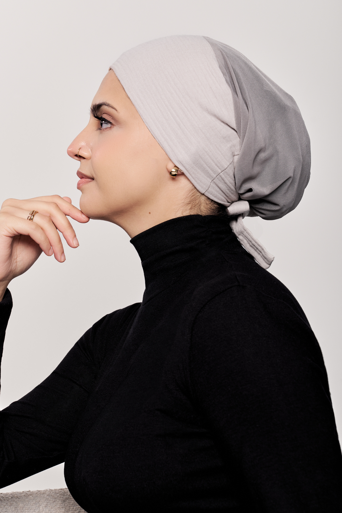 A woman in profile wearing a light underscarf and a black turtleneck top, posed against a neutral light gray background.