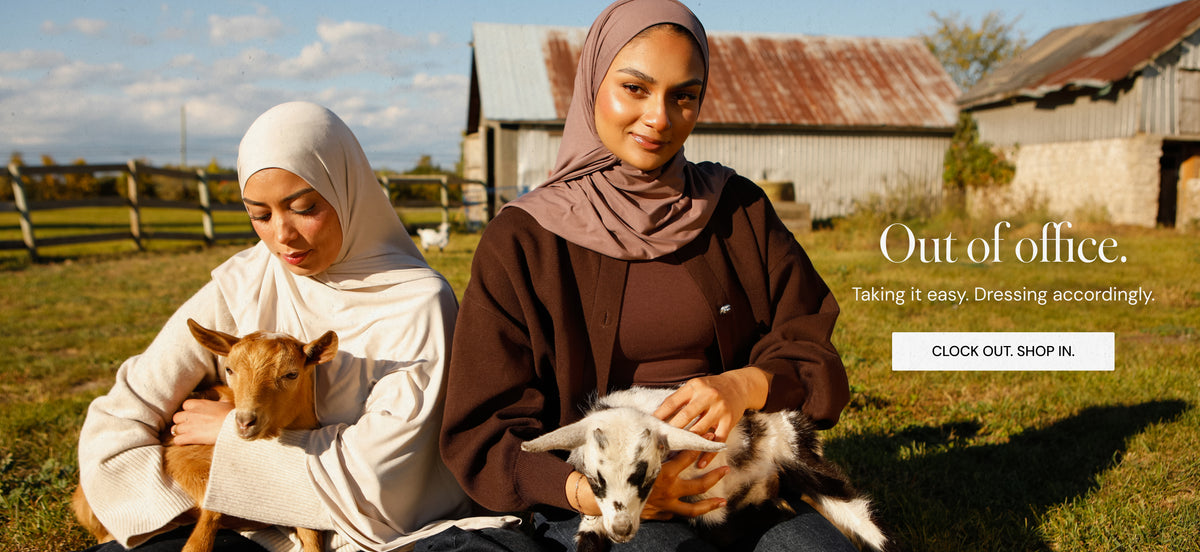 Two women wearing modest clothing  and bamboo jersey hijabs sit outdoors on a grassy field with goats, with rustic barns in the background. Text on the right reads 'Out of office. Taking its easy. Dress accordingly Click anywhere on the screen to view collection. 