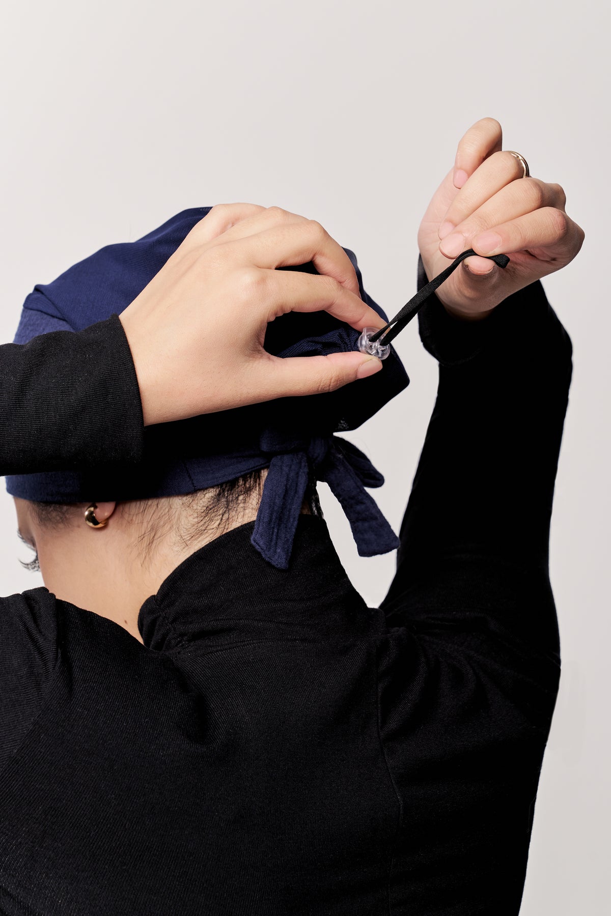 A person wearing a navy blue underscarf and a black long-sleeve top, shown from behind as they adjust the cap by an attached drawstring. 
